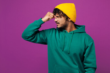 Young sad exhausted sick ill weary Indian man he wear green hoody yellow hat casual clothes keep eyes closed rub put hand on nose isolated on plain purple background studio portrait Lifestyle concept
