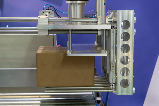Close-up view of modern heavy robotic arm manipulator moving large blank cardboard box on conveyer belt in industrial building. Soft focus. Copy space. Modern post storage and manufacturing theme.