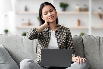 Upset asian young woman in casual suffering from neck pain while sitting on couch and using laptop,...