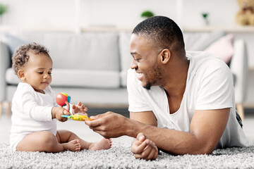 Obraz premium Development Activities With Babies. Portrait Of Caring Young Black Father Playing With Infant Child At Home, Happy African American Man Giving Toy To His Cute Adorable Toddler Baby And Smiling