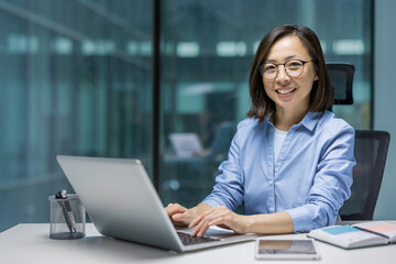 Portrait of a young successful Asian businesswoman, office worker smiling and looking at the camera, woman at workplace inside office working with laptop.