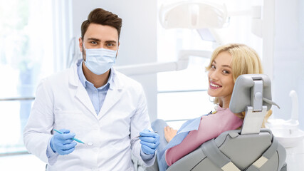 Dental clinic patient happy young blonde woman and male doctor in workwear holding dental tools, looking together at camera, dentist making check up for beautiful lady, modern dental cabinet interior