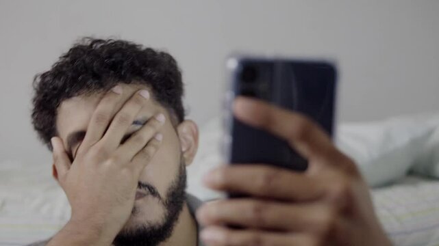 Hispanic man expressing frustration and covering mouth while holding smartphone