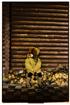 Child in warm coat sitting against log cabin with firewood stacked