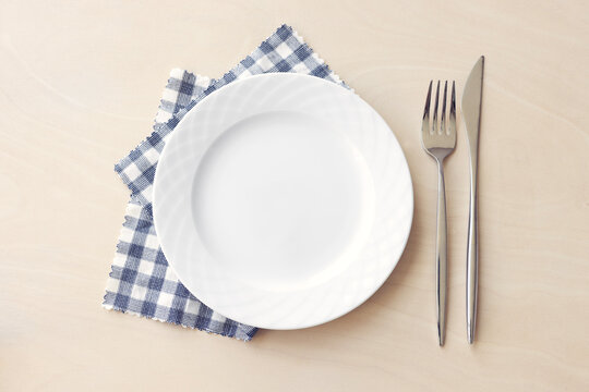 Empty white plate with blue fabric napkin, a fork and a knife on wooden table. Top view.