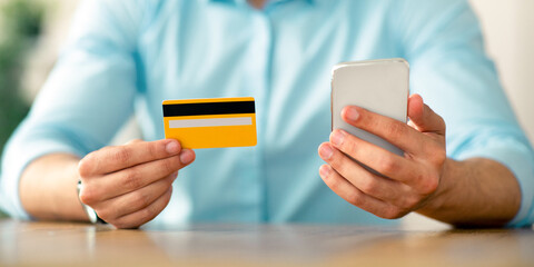 Online shopping concept. Unrecognizable Arab guy holding credit card and cellphone, making e-payment, ordering products on internet, purchasing goods on web at office, croped view