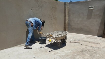 Homem de roupa azul e chapéu preto, trabalhando na construção do muro, com carriola de cimento e balde com água ao lado.