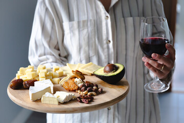 A woman holding a wooden platter with a variety of cheeses, nuts, dried fruits, and half an avocado and glass of wine