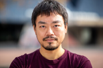 Close-up portrait of an Asian man in maroon t-shirt outdoors with focused expression