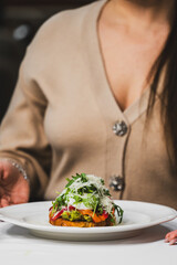 A beautifully plated gourmet dish featuring colorful vegetables and garnished with fresh herbs on a white plate. A well-dressed individual is holding the plate, enhancing the dining experience.