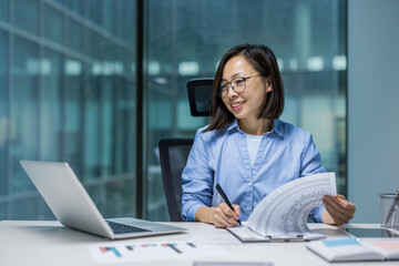 Joyful and successful Asian woman doing paperwork inside the office, businesswoman financier...
