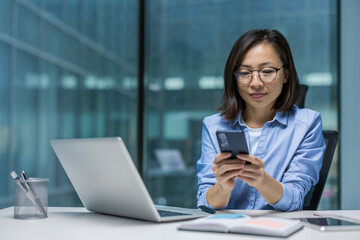 Serious and focused Asian woman using an app on her smartphone, businesswoman with phone in hands working sitting inside office with laptop.