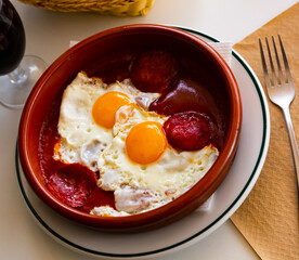 Popular dish of spanish cuisine, fried eggs with chorizo sausages and tomato paste served on plate in cafe