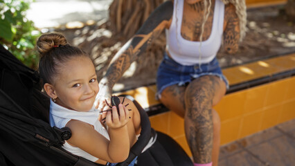 Woman with tattoos sitting on a park bench with young girl in stroller enjoying a cookie outside, embodying urban family love in a town setting