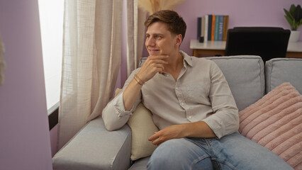 Obraz premium Young man sitting on a couch in a cozy living room with a thoughtful expression on his face, light from the window illuminating the interior.