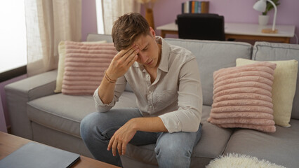 Young man sitting in a living room with a pensive expression, wearing casual clothing, surrounded...