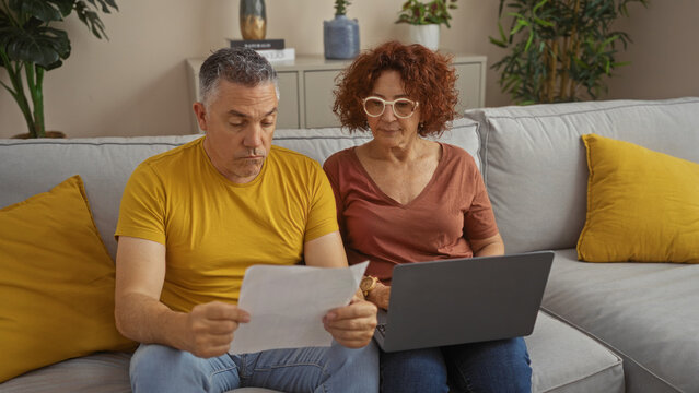 Middle-aged couple sitting together on a living room sofa reviewing documents and using a laptop showcasing a home interior setting