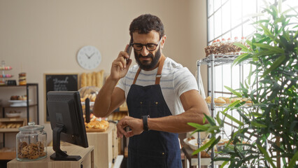 Young hispanic man in a bakery wearing an apron, looking at his watch, talking on the phone,...