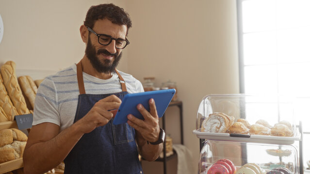 Young hispanic man using tablet in bakery wearing apron surrounded by pastries showcasing modern technology in traditional setting