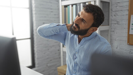 Fototapeta premium Young man with beard and blue shirt stretching his neck in a bright office, surrounded by books and documents, suggesting a professional and focused work environment.