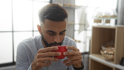 Handsome young hispanic man with a beard enjoys a coffee in a cozy bakery shop, highlighting his...