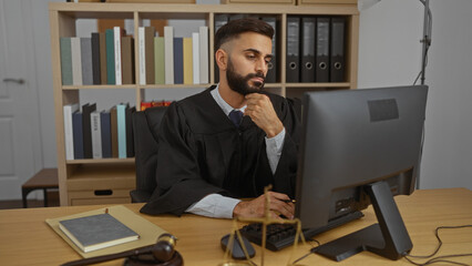 Young hispanic man in judge robe working at computer in office with bookshelf in background and...