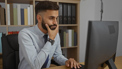 Handsome, hispanic, man with, beard working, in an, office while, sitting at, a desk, indoors.