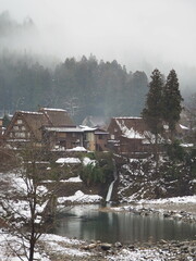 Snow-covered Gassho-style houses in Shirakawa-go, Japan, surrounded by misty mountains and a calm river.