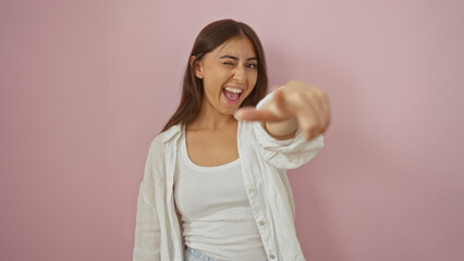 Beautiful brunette woman pointing and winking over isolated pink background exuding joy and...