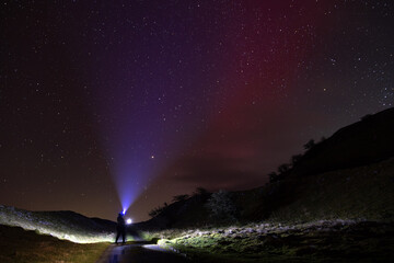 Aurora Boralis, The Northern lights in Wales 1st January 2025. The vibrant night sky glows with shades of red and purple. In the foreground, a figure stands holding a torch light.