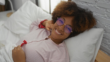 Woman relaxing on bed in cozy bedroom, african american female smiling with curly hair and glasses,...
