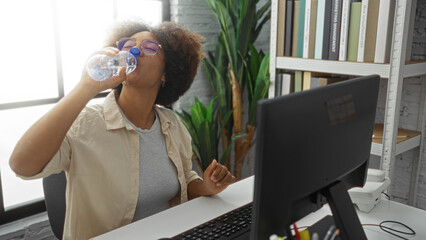 Woman drinking water in office room with curly hair seated at desk indoor environment displaying work setting and hydration focus.