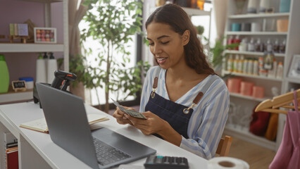 Young woman working in a home decor store counting money at a desk with a laptop and various decorations on shelves in the background.