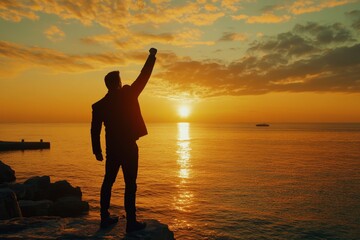 Fototapeta premium Silhouette of a celebrating businessman with raised fist at sunset by the sea, capturing success