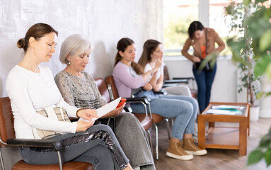 Adult woman in casual clothes looks through documents while waiting for her turn while sitting on chair in reception