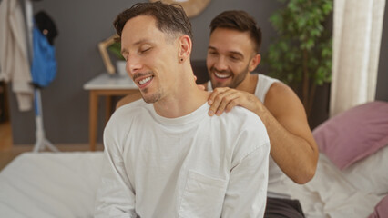 Men in a bedroom with one giving the other a back massage while sitting on a bed and smiling