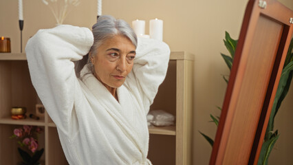 Woman grooming herself in a serene spa setting, wearing a white robe, looking in the mirror with candles and plants in the background, showcasing relaxation and self-care.