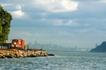 Red freight train with lights rounding the bend of the Hudson River with skyline of NYC in background