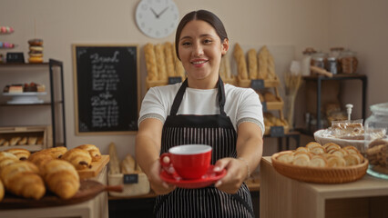 Young brunette woman with a happy smile in a bakery holding a red cup amidst fresh pastries and breads with a cozy interior