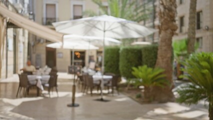Defocused outdoor cafe scene with empty tables, sun umbrellas, greenery, and blurred architecture in background on a sunny day.