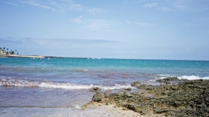 Blurred beach scene with defocused view of ocean, waves, and rocky shoreline under a clear sky.