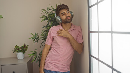 Young hispanic man looking through window at home wearing headphones indoors