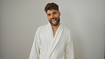 Handsome young hispanic man with beard wearing white robe and sticking out tongue while standing isolated over white wall background