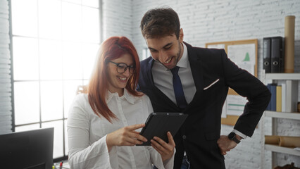Woman and man executives in office sharing ideas on tablet smiling indoors professional setting business workplace collaboration.