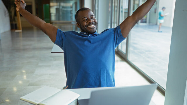 Smiling man stretching in an office, sitting at his desk with a laptop, capturing a moment of relaxation and joy in a modern workplace.