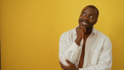 Young man thinking with a smile over isolated yellow background, showcasing an african american adult male with thoughtful expression and casual attire