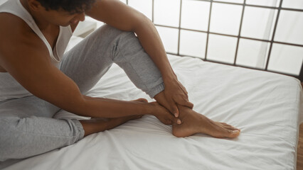 Young man sitting on bed in bedroom adjusting his ankle wearing casual clothes