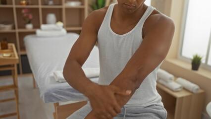 Young man in a spa wearing casual gym clothes, seated in a tranquil beauty salon, with a modern wellness room backdrop featuring a massage table and organized shelves.