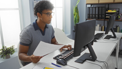 Young man working in an office, holding papers and typing on a computer, in a bright indoor workplace with a modern setup and various office supplies around.