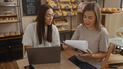 Women working together in a bakery, discussing business documents with a laptop on the table, surrounded by bread and pastries, highlighting their collaboration in a cozy indoor shop.